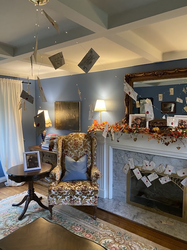 A cozy living room decorated for a Harry Potter themed party, featuring floating envelopes, a floral armchair, and a fireplace adorned with fall decorations. Pictures of the Dursley's on the mantle with Dudley, Petunia, and Vernon Dursley.